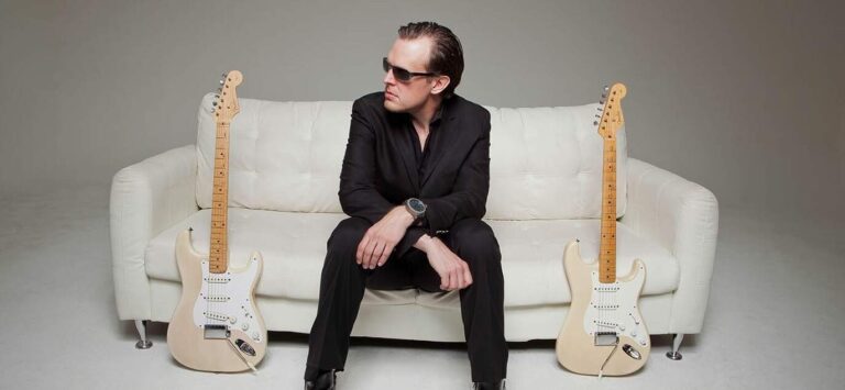 Joe Bonamassa sentado no sofá com duas guitarras Fender Stratocaster