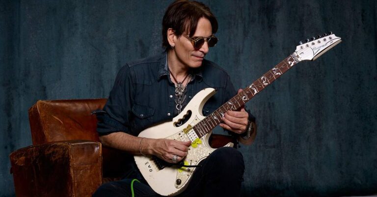 Steve Vai tocando guitarra sentado em um sofá