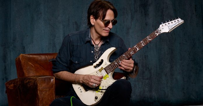 steve-vai Steve Vai tocando guitarra sentado em um sofá
